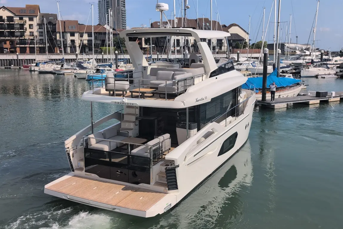 A luxurious white yacht named "Navetta 53" docked in a marina, with boats and waterfront buildings visible.